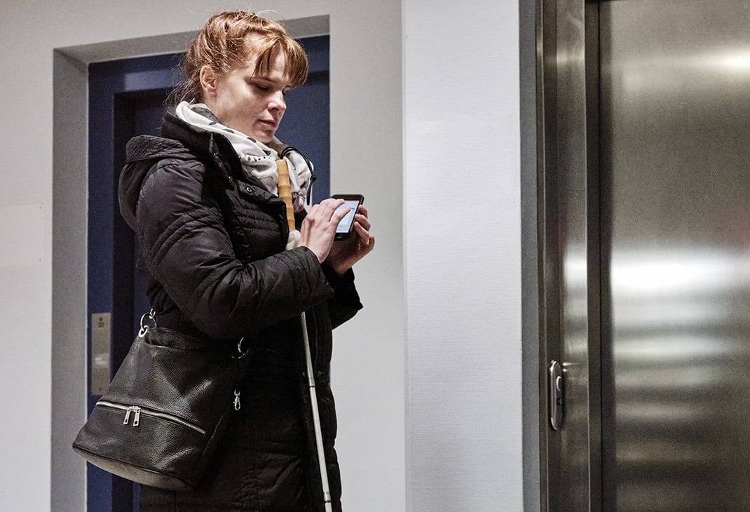 A woman who is blind using the BlindSquare application on her mobile phone in front of an elevator, white cane in hand. 
